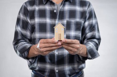 Bir adam ahşap bir ev modelini elinde tutuyor, şık bir kareli gömlek giyiyor. Bu resim ev sahipliğini ve gelecek kurma hayallerini simgeliyor..