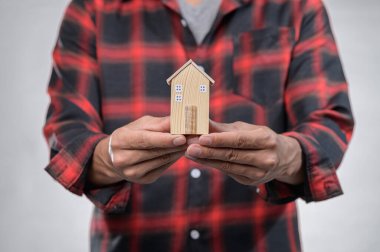 Sıradan fanila gömlekli bir insan, ev sahipliği ve sıcaklığı sembolize eden minyatür ahşap bir ev modeli taşıyor. Yaşam tarzı ve mimari temalar için ideal.