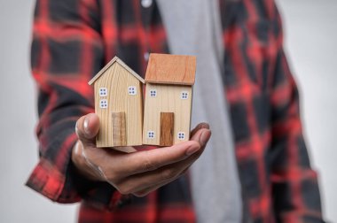 Flanel gömlek giyen bir insan, yaratıcı bir tasarım sergileyen iki küçük ahşap ev taşır. Bu resim ev sahipliğini, işçiliği ve inşaatın neşesini simgeliyor..