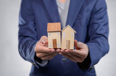 Bir iş adamı, emlak yatırımı ve ev sahipliği sembolize ederek iki küçük ahşap model ev sergiliyor. Resim finans ve tasarım kavramlarını iletir.