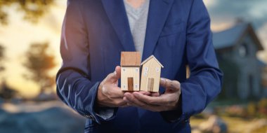 Elinde iki model ev tutan kişi emlak piyasasının satın alma, satma ve kiralama yönlerini sembolize ediyor, konut alım-satım işlemlerini vurguluyor.