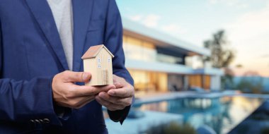 Profesyonel bir birey elinde minyatür bir ev tutuyor ve günümüzde konut satın almanın çeşitli yönlerini sembolize ediyor.