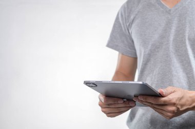 Gri tişörtlü bir kişi elinde bir tablet tutuyor ve modern teknoloji kullanımını vurguluyor. Işık arkaplanı cihaz ve ellere odaklanmayı arttırır, yaşam tarzı temaları için mükemmeldir..