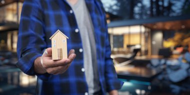 Bir kişi ev alıp satma kavramını sembolize eden bir model ev tutuyor. Bu görüntü, modern bir ortamda gayrimenkul işlemlerinin özünü yansıtıyor..