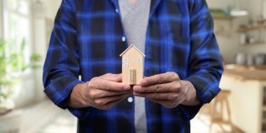 Bir kişi, emlak alım ve satım sürecini sembolize eden minyatür bir ev modeli tutuyor. Bu resim ev sahipliği ve kiralama seçeneklerinin temalarını yakalar.