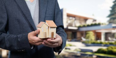 Takım elbiseli bir profesyonel ahşap model evler tutuyor, modern şehir ortamında gayrimenkul satışlarını ve emlak yatırımlarını sembolize ediyor..