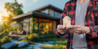 Kiralık konutları alıp satma kavramını temsil eden minyatür bir ev modelini elinde bulunduran bir kişi, güzel bir açık hava arka planına sahip..