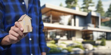 Bir kişi emlak alım ve satım sürecini sembolize eden ahşap bir ev modeli tutuyor. Arka plandaki modern mülk mülkiyet kavramını geliştirir..