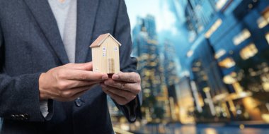 Bir işadamı şehir ortamında örnek bir ev tutuyor, gayrimenkul satışları ve yatırım fırsatlarını sembolize ediyor, modern şehir yaşamını ve profesyonelliğini gözler önüne seriyor.