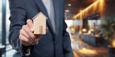 Zarif bir iç mekanda ahşap bir ev modeli tutan bir işadamı, emlak sektöründeki ev satışlarını sembolize ediyor. Gayrimenkul pazarlama veya iş kavramları için ideal.
