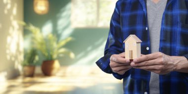 Bir kişinin minyatür bir ev modeli tuttuğunu gösteren kavramsal görüntü, parlak ve davetkar bir iç mekanda ev alıp satışı ve kiralamayı sembolize ediyor..