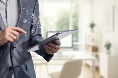 Modern ofis ortamında bir tabletle uğraşan bir işadamı, dijital arayüzler ve elementlerle teknolojik etkileşim sergiliyor. Yenilik ve profesyonellik temaları için mükemmel.
