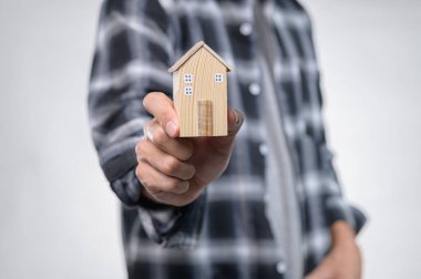 Bir kişi, ev sahipliği ve yeni başlangıçları simgeleyen küçük ahşap bir ev modeli sergiliyor. Arka plan hafifçe bulanık, modelimize odaklanıyoruz..