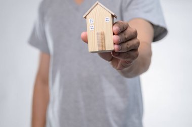 Gri tişörtlü bir adam küçük tahta bir model evi tutuyor, yumuşak bir ışık arka planına dayanıyor. Bu resim ev sahipliği, tasarım ve inşaatı sembolize eder.