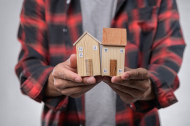 Ekose gömlekli bir insan, iki adet el yapımı ahşap ev modeli taşır. Yaratıcılık ve zanaatkarlık sergiler. Ev ve sıcaklık içeren tasarımlar için mükemmel..