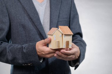 Takım elbiseli profesyonel bir birey, emlak yatırımı ve mülkiyeti sembolize eden iki ahşap model ev tutuyor. Ayarlama zarafet ve hırs taşır..