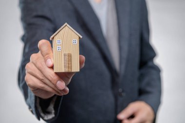 İş kıyafetleri içinde bir kişi, emlak yatırımını ve konut kavramlarını sembolize eden ahşap bir ev modeli taşıyor. Finans veya mimari promosyonlar için ideal.