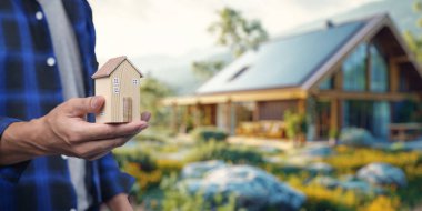 Güzel bir evin önünde küçük bir model evi tutan birini gösteren kavramsal bir görüntü. Bu, gayrimenkul alıp satma süreçlerini sembolize ediyor..