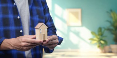 Ahşap bir model evi elinde tutan kişi, emlak alıp satma kavramını temsil ediyor. Bu resim gayrimenkul yatırımının ve ev sahipliğinin özünü aktarıyor..