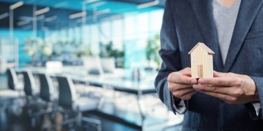 Modern bir ofiste, elinde küçük bir model ev bulunan bir iş adamı gayrimenkul satışlarını ve ev yatırımlarını sembolize ediyor. Bu resim iş ve konut piyasaları arasındaki ilişkiyi vurgular.