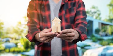 Bir kişi, konut kiralama ve satma kavramını temsil eden minyatür bir ev modeli tutuyor. Arka planda, yatırım fırsatlarını sembolize eden sakin bir doğa sahnesi yer alıyor..