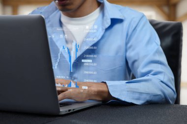 Bir finans uzmanı dizüstü bilgisayar kullanarak çeşitli hisse senetlerini analiz eder ve ekranda veri görselleştirmesini gösterir. Çevre, pazar eğilimleri ve mali anlayışlara odaklanmış bir çalışma alanı anlamına gelmektedir.