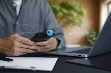 Mobil bir cihaz ve dizüstü bilgisayar kullanan, soyut simgelerle dijital güvenlik tasvir eden, modern bir çalışma alanında teknolojiyi ve mahremiyeti vurgulayan bir kişinin yakın plan görüntüsü.