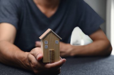 Ahşap bir ev modelini günlük kıyafetle tutan kişi, emlak ticaretini ve emlak yatırımını sembolize ederek konut piyasasının ve finansal fırsatların özünü yansıtıyor..