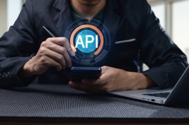 Profesyonel bir adam, iş ortamında modern programlama arayüzleri ve dijital teknolojiyi temsil eden API sembolü gösteren mobil bir cihazla etkileşim halindedir..