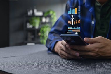 Akıllı telefon kullanan bir kişi sürdürülebilirlik ve yenilenebilir enerji ile ilgili görsel veriler sergiler, eko-dost uygulamalardaki teknolojik gelişmeleri vurgular ve izleme.