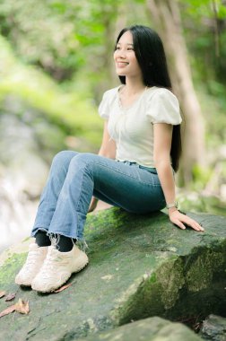 Genç bir kadın yemyeşil sularla çevrili çarpıcı şelalelerin yakınındaki bir kayanın üzerinde huzur içinde oturuyor. Parlak gülüşü doğadaki huzur ve neşeyi yansıtıyor..