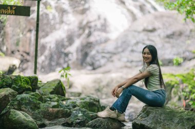 Genç bir kadın, yemyeşil ve sakin sularla çevrili güzel Kuzey ve Güney Jetkot Şelaleleri yakınlarındaki bir kayanın üzerinde huzur dolu bir anın tadını çıkarıyor..