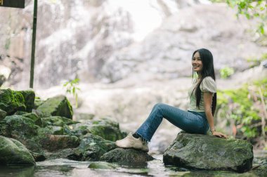 Genç bir kadın canlı yeşilliklerle çevrili yumuşak bir şelalenin yanındaki bir kayanın üzerinde neşeyle oturuyor. Bu sakin açık hava sahnesi rahatlamanın ve doğanın güzelliğini yansıtıyor..