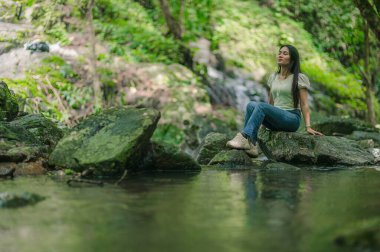 Huzurlu bir kadın, verimli yeşillikler ve doğanın huzurlu sesleri ile çevrili güzel şelalelerde kayaların üzerinde oturmaktan zevk alır. Bu resim huzur ve rahatlığı huzurlu bir ortamda yakalar..