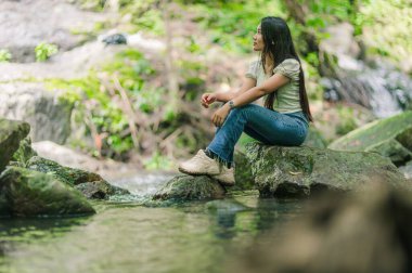 Nehir kenarındaki sakin bir anda, bir kadın yemyeşil şelalelerle çevrili şelalenin yakınındaki doğanın güzelliğinin tadını çıkararak dinlenmek için huzurlu bir atmosfer yaratıyor..