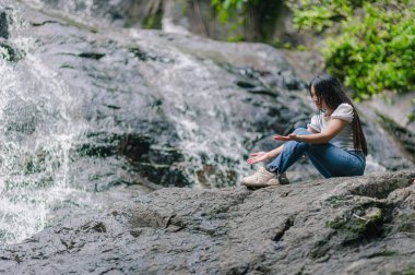 Genç bir kadın Kuzey Jetkot Şelaleleri 'nin oradaki bir kayanın üzerinde huzur içinde oturuyor. Doğanın güzelliğinin ve akan suyun rahatlatıcı seslerinin tadını çıkarıyor. Rahatlama temaları için mükemmel.