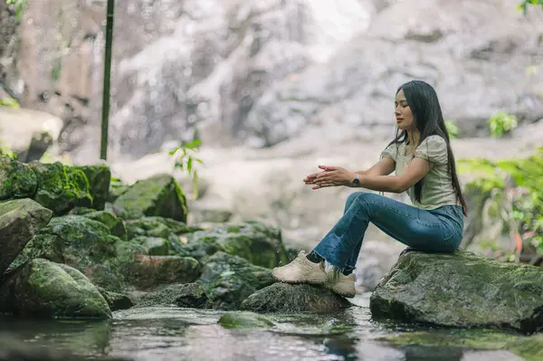 Bir kadın şelalenin yanında huzur içinde oturur, doğanın huzurlu güzelliğinin tadını çıkarır. Etrafı yemyeşil ve kayalarla çevrili, sakin sulara yansıyor..