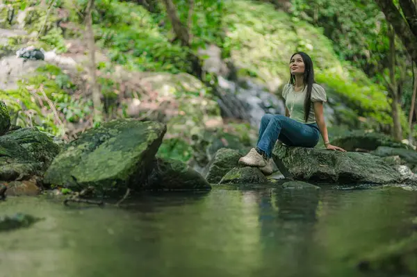 Huzurlu bir kadın yemyeşil ve pürüzsüz kayalarla çevrili bir şelalenin yanında oturur. Bu barışçıl ortam, doğayla bir huzur ve bağlantı anı yakalıyor..
