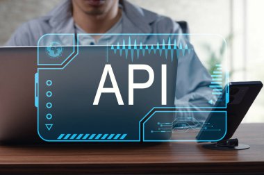 Odaklanmış bir birey, bir dizüstü bilgisayar ve akıllı telefon üzerinde API grafiği barındıran teknolojiyle etkileşim halindedir. Bu resim modern programlamayı ve dijital yeniliği sembolize eder..