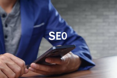 Bir iş adamı akıllı telefonuyla çalışır, arama motoru optimizasyonu stratejilerine odaklanır. Bu görüntü modern pazarlama çabalarını temsil ediyor.