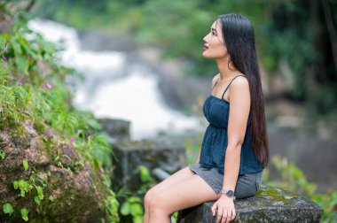 Genç bir kadın, yemyeşil ve çağlayan şelalelerle çevrili gür bir ormanda huzurlu bir derenin kenarında huzur içinde oturur ve doğadaki huzurlu bir anı yakalar..