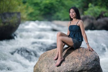 Doğanın güzelliğini, akan şelalelerin yanında bir kayanın üzerinde oturan genç bir kadının bu huzurlu görüntüsüyle kucaklayın. Yeşillik ve canlı nehirler arasında sükuneti hissedin..