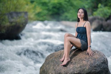 Doğanın dingin güzelliğini, akarsuları ve şelaleleri yemyeşil bir ormanda deneyin. Genç bir kadın, bir kayanın üzerinde huzur dolu bir anın tadını çıkarıyor. Manzaranın içinde..