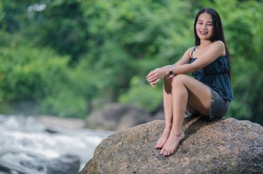 Bereketli bir ormandaki bir derenin kenarındaki kayanın üzerinde oturan bir kadının huzurlu görüntüsü. Yeşillik ve akan suyla çevrili doğanın güzelliğinden zevk alır. Rahatlamak ve macera yaşamak için mükemmeldir..