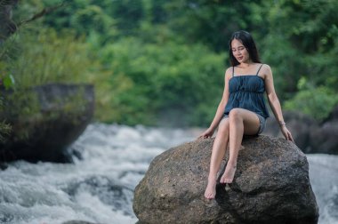 Canlı bir ormandaki canlı bir şelalenin yanındaki bir kayanın üzerinde oturan bir insanın sakin sahnesinin tadını çıkarın. Doğanın güzelliği rahatlamayı ve keşfi davet eder..