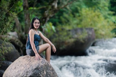 Yoğun, yeşil bir ormandaki akarsuyun yanındaki bir kayanın üzerinde dinlenen bir kadın. Sahne, şelalelerin güzelliği artırdığı doğanın sükunetini yakalıyor..