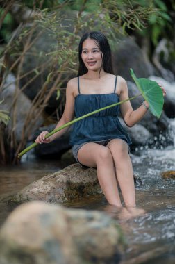 Bir kadın yemyeşil yemyeşil bir dere kenarında huzurlu bir anın tadını çıkarır. Güneş ışığı, huzurlu ortamın doğal güzelliğini arttıran sıcak bir parıltı yayar..