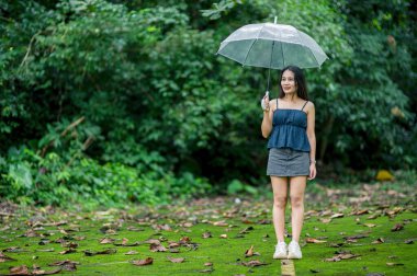 Canlı bir ormanda elinde temiz bir şemsiyeyle duran bir kadın. Yeşillik ve dağınık yapraklarla çevrili, doğadaki maceranın özünü somutlaştırıyor..