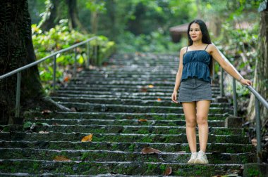 Genç bir kadın canlı bir ormandaki taş basamaklarda zarif bir şekilde duruyor. Doğanın güzelliğini ve yemyeşil ortamda yapılan keşiflerin neşesini gözler önüne seriyor..