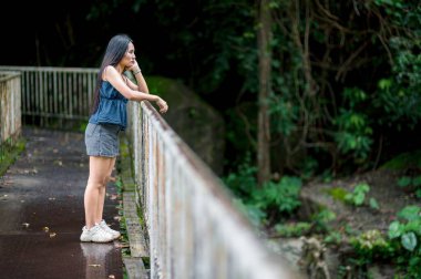 Genç bir kadın düşünceli bir şekilde bir orman yolunda duruyor, doğanın güzelliğine bakıyor. Bu sakin sahne gezinin ve açık havada keşiflerin özünü yakalar..